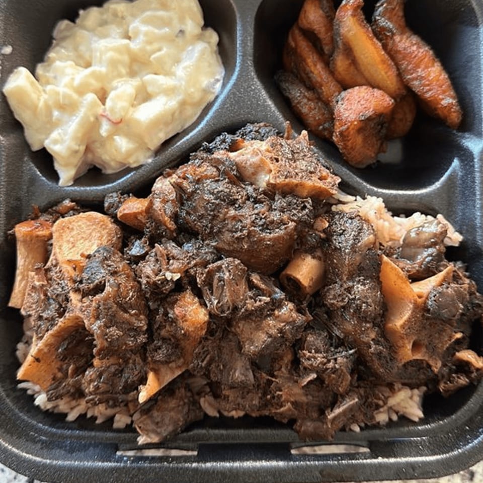 Jamaican oxtail with rice, macaroni salad, and fried plantains in a black takeout container.