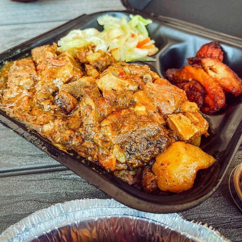 A Jamaican oxtail stew with cabbage, carrots, and fried plantains in a black takeout container.
