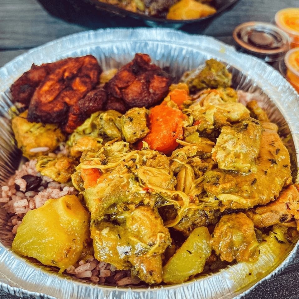 A Jamaican meal in a foil container with curry chicken, rice and peas, and fried plantains.