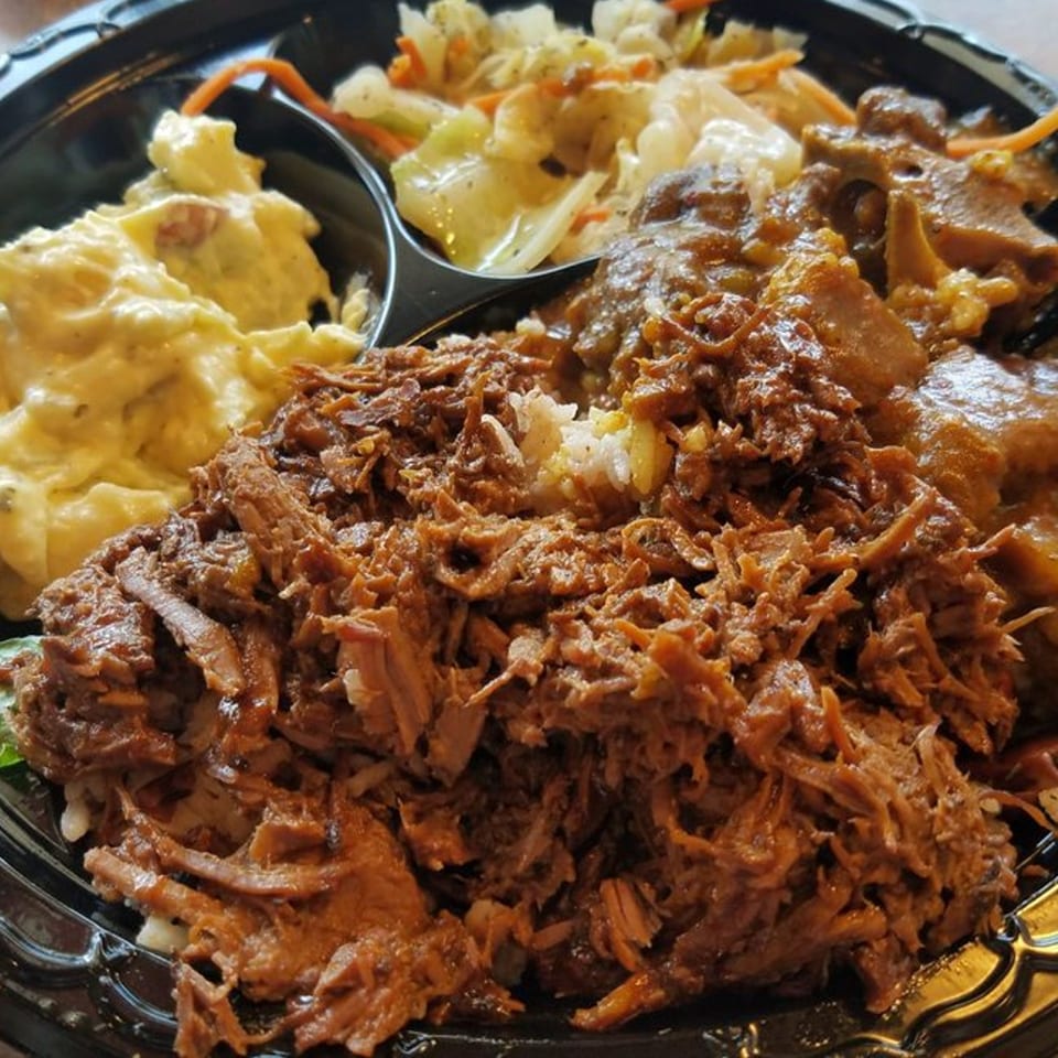 A Jamaican meal with pulled pork, potato salad, cabbage, and oxtail in a black divided plate.